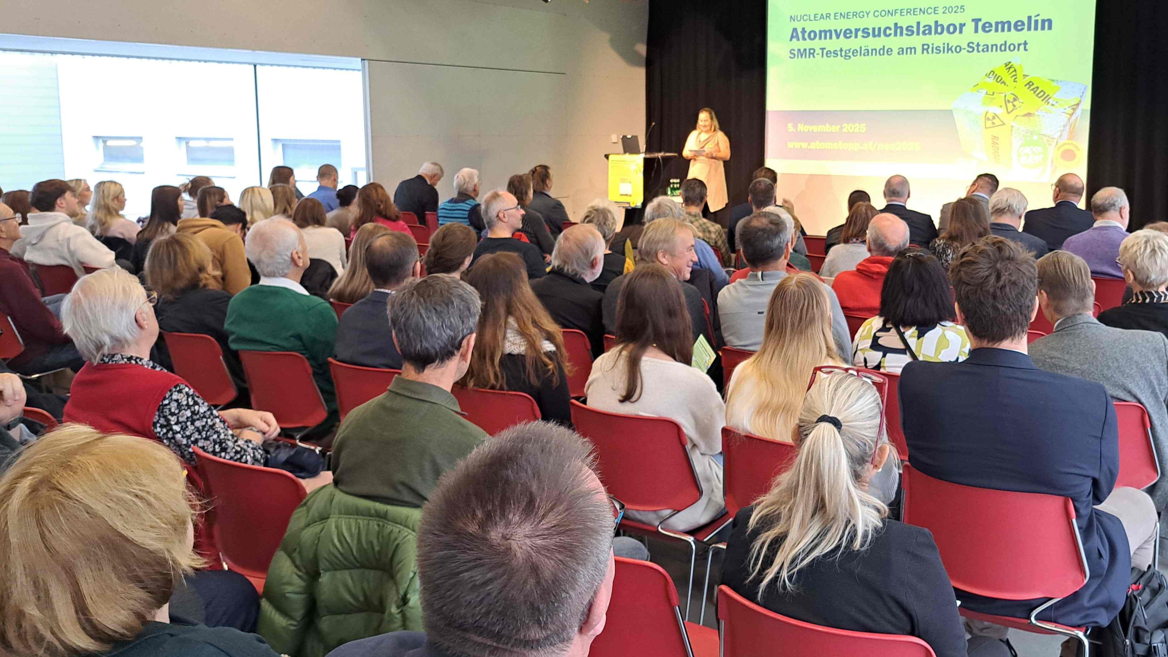 Freundlich lächelnde Moderatorin Weinbauer spricht auf der Bühne zum Publikum im Bildvordergrund, etwa 50 Menschen unterschiedlichen Alters. Im Hintergrund die Präsentationsleinwand mit Textfolie: Nuclear Energy Conference Atomversuchslabor Temelín - SMR-Testgelände am Risiko-Standort